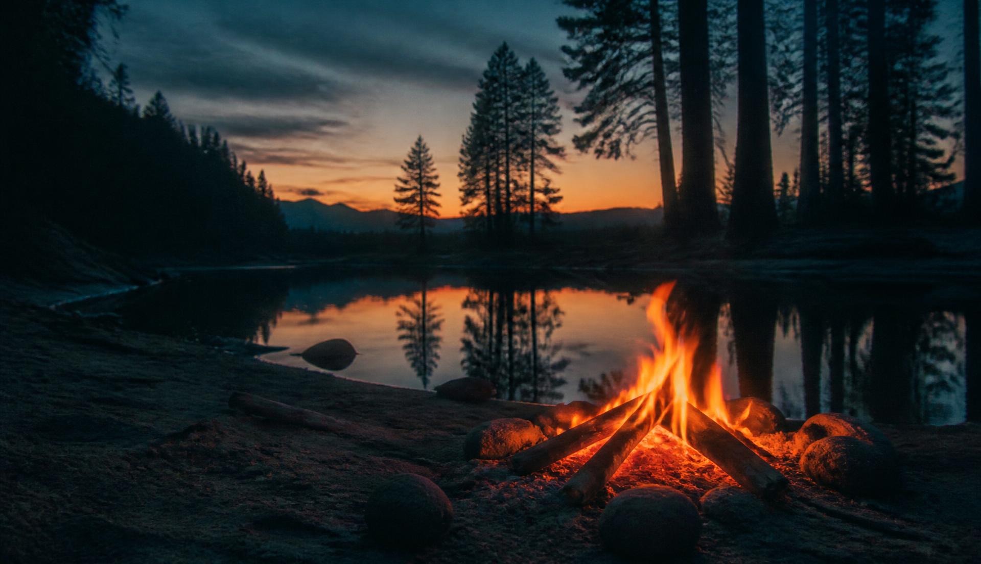 A fire pit burning in the sand beside the pond at dusk